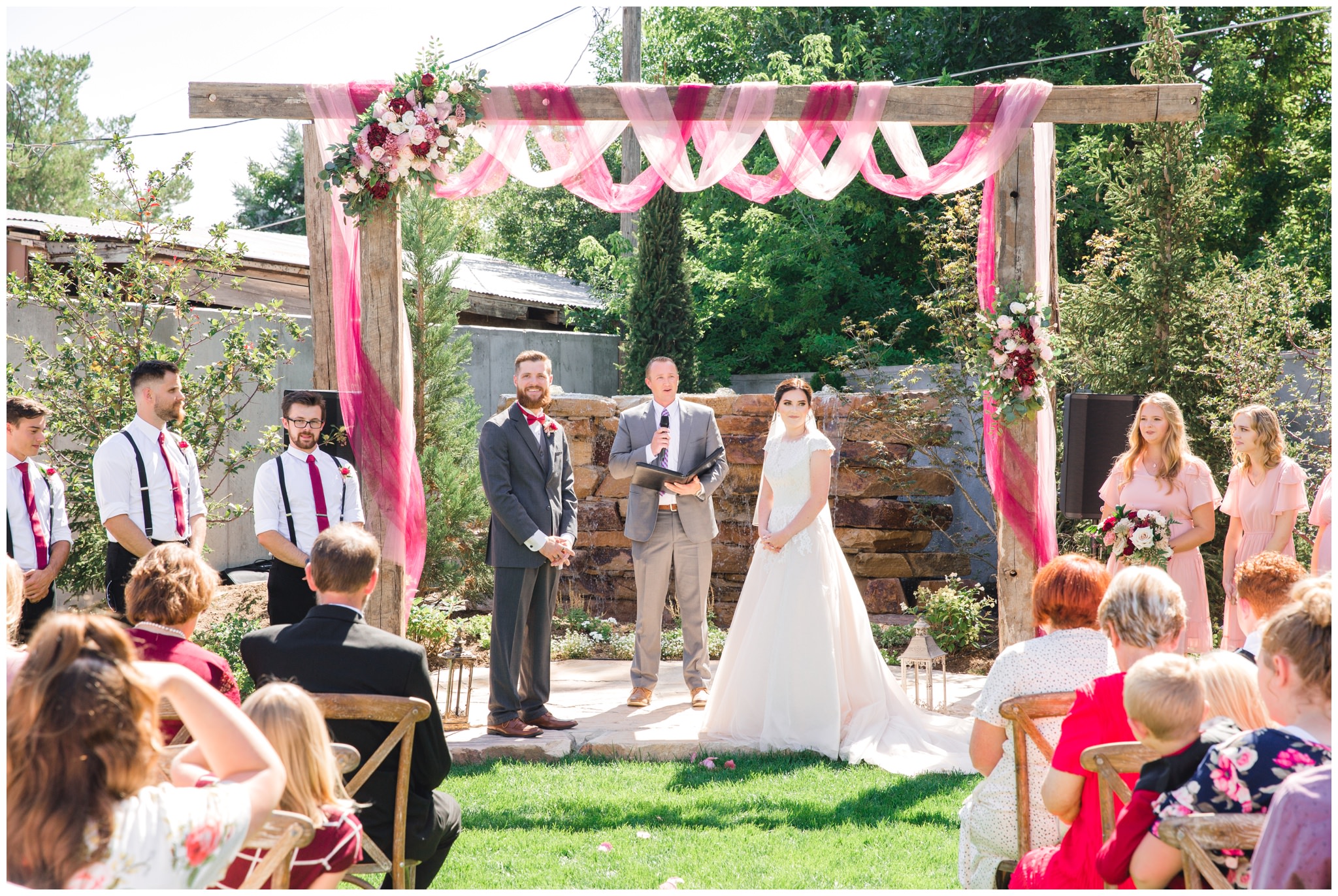 Summer wedding ceremony at the Wild Oak Venue in Lindon, Utah. Burgundy floral arrangement on arch above bride and groom.